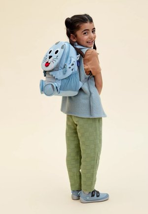 Light blue backpack with animal face design, plush fabric, round ears, prominent eyes, and a playful tongue, worn by child in green textured pants.