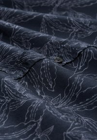 Donkere stoffen shirt met een witte botanische print. Inclusief een knoopdetail op de kraag. Glad textuur met zichtbare gevouwen lagen.