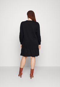 Vestido preto de mangas longas com bainha volutuosa, feito de um tecido suave, combinado com botas de tornozelo castanhas. A vista de trás destaca a textura suave e o design.