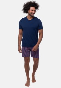Marineblauwe v-hals t-shirt gecombineerd met rood en blauw gestreepte zwemshorts. De stof lijkt lichtgewicht te zijn. Blootsvoets, staand op een effen achtergrond.