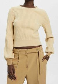 Pull en maille côtelée crème avec un col bateau et des manches longues bouffantes, associé à un pantalon taille haute de couleur fauve avec une ceinture à la taille.