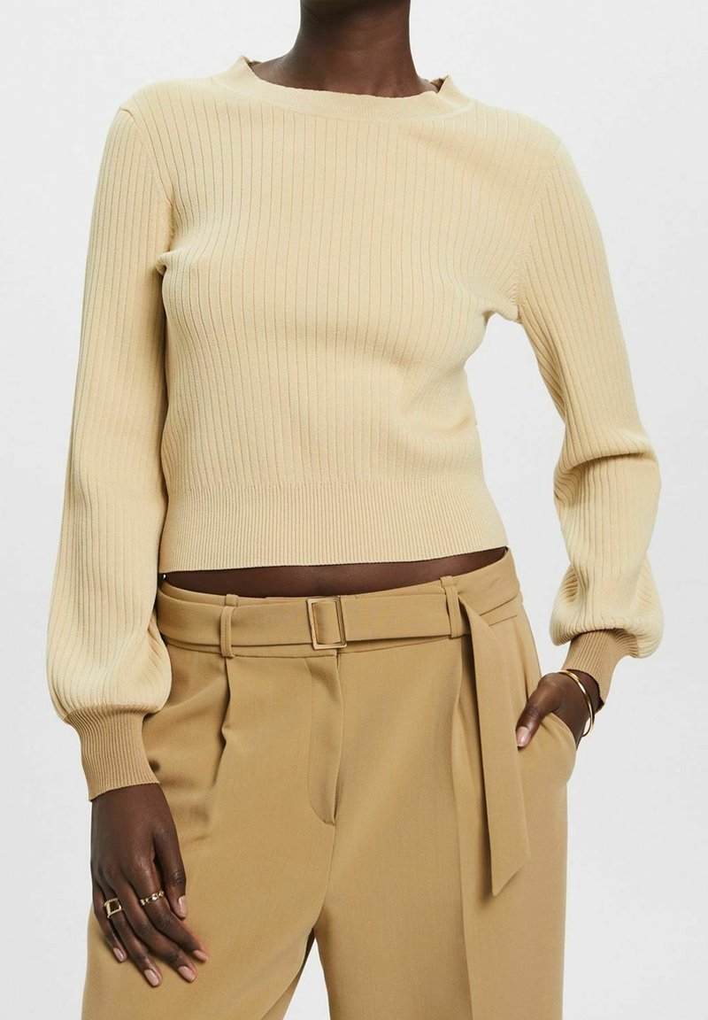 Pull en maille côtelée crème avec un col bateau et des manches longues bouffantes, associé à un pantalon taille haute de couleur fauve avec une ceinture à la taille.