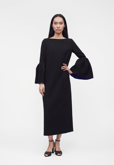 Γυναίκα με μαύρο μακρύ φόρεμα με φαρδιά μανίκια καμπάνα και μαύρα ψηλοτάκουνα σανδάλια, στέκεται μπροστά σε απλό ανοιχτό φόντο.