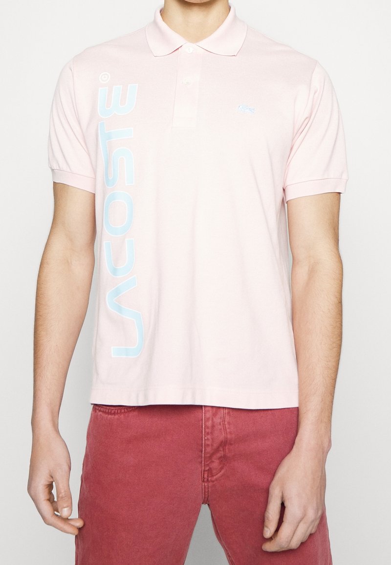 Homme portant un polo Lacoste rose clair avec un logo vertical blanc sur le côté gauche, associé à un pantalon rouge, debout devant un fond uni.
