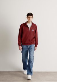 Βουργουνδί κόκκινο pullover με κουμπί στο μισό, παχύ ύφασμα, λευκό λογότυπο στο στήθος, συνδυασμένο με ανοιχτό μπλε τζιν και λευκά αθλητικά παπούτσια, απαλός, άνετος κόψιμο.