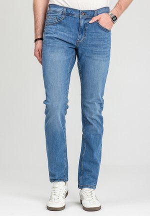 Mężczyzna w jasnoniebieskich, dopasowanych dżinsach i białych trampkach, z jedną ręką w kieszeni, zegarek na lewej ręce, stojący na szarym tle.