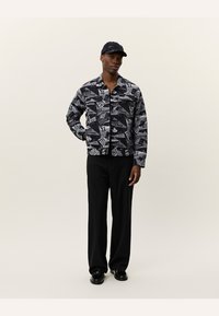 Camicia a motivi neri e grigi con vari disegni grafici, design con bottoni e maniche lunghe, abbinata a pantaloni e scarpe nere.