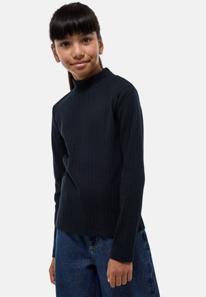 Pull à col roulé noir côtelé à manches longues en tissu texturé, associé à un jean en denim bleu à jambes larges. Le modèle se tient devant un fond neutre.