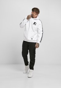 Hanorac alb cu glugă, cu un buzunar frontal, logo negru pe piept și accente cu model negru pe mâneci. Purtați cu pantaloni cargo negri și adidași albi.