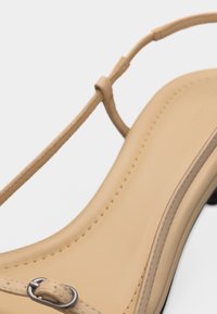 Μπεζ δερμάτινο σανδάλι με λεπτό λουράκι στον αστράγαλο και μικρή ασημί αγκράφα, που δείχνει το ραμμένο πάτο και την λεπτομέρεια του χαμηλού τακουνιού.