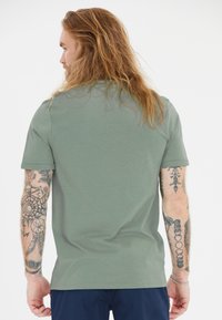 Vihreä puuvillapaita lyhyillä hihoilla, pyöreällä kaula-aukolla, sileä rakenne, henkilö, jolla on tatuointeja, näkyy takaa.