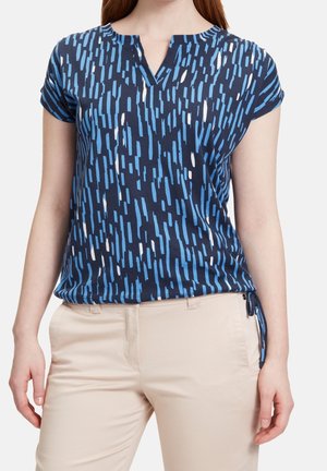 Kobieta ubrana w granatowy top z krótkimi rękawami z nieregularnymi pionowymi paskami w jasnoniebieskim i białym kolorze, zestawiony z beżowymi spodniami.