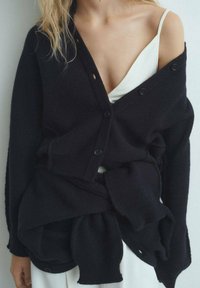 Cardigan negro de punto con un corte holgado, que presenta botones grandes y tela superpuesta. Llevado sobre un camisón blanco, mostrando una textura suave.