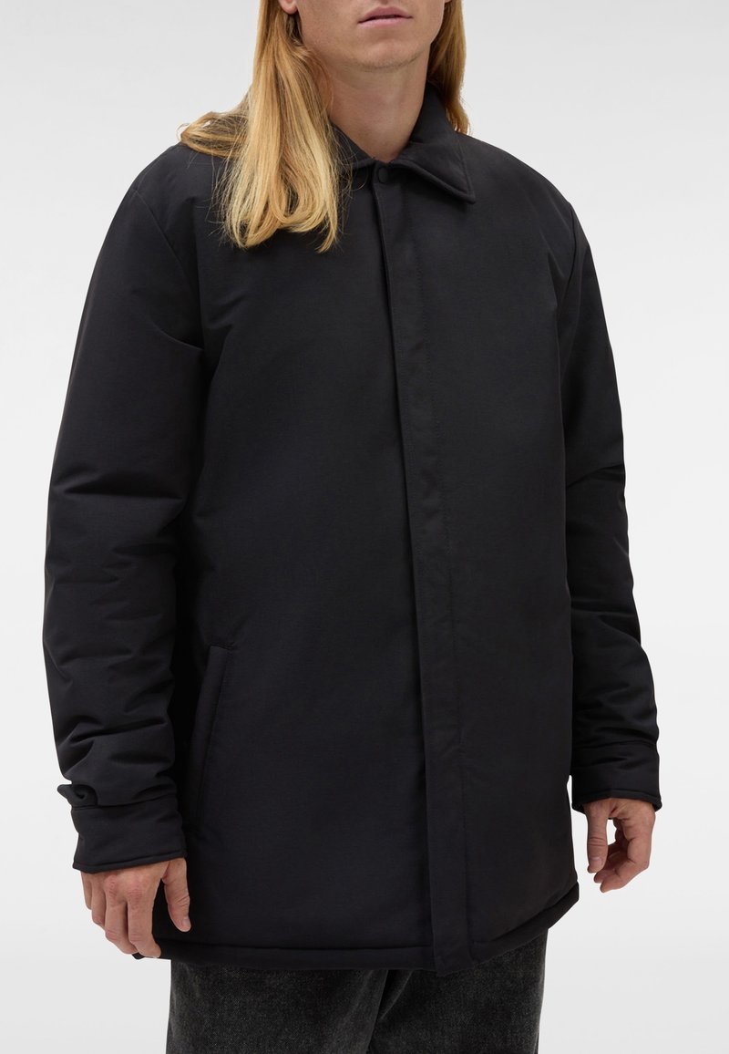 Zwarte, geïsoleerde jas met een afgeronde kraag, knoopsluiting en zijzakken. Soepele textuur, design met middellange lengte en licht oversized pasvorm.