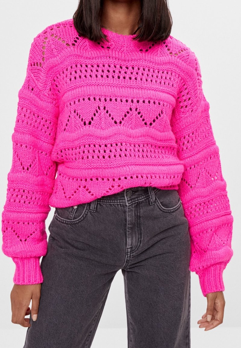 Różowy dzianinowy sweter z teksturowanym, ażurowym wzorem, okrągłym dekoltem i ściągaczami na rękawach, zestawiony z ciemnoszarymi, wysokim stanem jeansami.