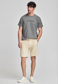 Camiseta gris de manga corta con el texto "POLO CLUB", combinada con pantalones cortos beige. Zapatillas blancas completan el atuendo. Diseño simple y casual.