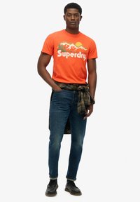 Orange bomulds T-shirt med bjerggrafik og "Superdry" logo, parret med mørkeblå jeans og en ternet skjorte bundet rundt om livet. Sorte sko.