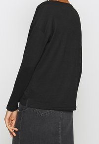 Haut noir à manches longues avec un col rond, une coupe décontractée et une texture lisse. Dispose d'une fente latérale à l'ourlet pour faciliter les mouvements.