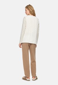 Woman wearing a cream cable knit vest over a long-sleeve top, beige knit pants, and brown shearling-lined moccasin slippers, viewed from behind.