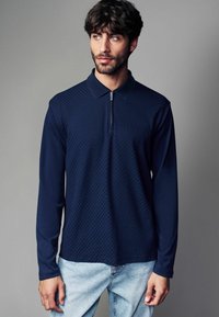 Polo cu mânecă lungă de culoare bleumarin, cu un model texturat în diamante, închidere cu fermoar la jumătate și guler clasic, asortat cu blugi de culoare albastru deschis.