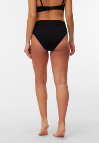 Γυναίκα που στέκεται ξυπόλυτη, φορώντας μαύρο σουτιέν και μαύρο εσώρουχο ψηλόμεσο, εμφανιζόμε νη από πίσω μπροστά σε απλό λευκό φόντο.