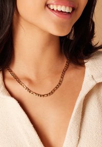 Gouden ketting met in elkaar grijpende schakels, gemiddelde dikte, glanzende afwerking, gedragen dicht bij de nek. Zacht wit stoffen kledingstuk op de achtergrond.