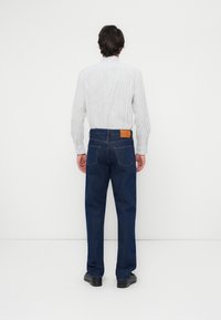Jeans de mezclilla azul oscuro con corte recto, que presentan un parche de cuero marrón en la parte trasera de la cinturilla y costuras de contraste. Combinados con una camisa a rayas.