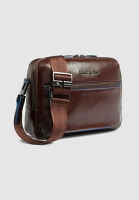 Borsa a tracolla in pelle marrone con una texture liscia, cuciture di accento blu e una tracolla marrone staccabile con fibbia in metallo.