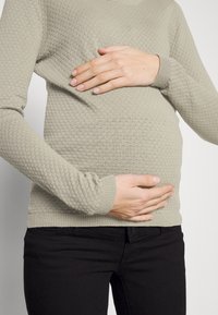 Pull en maille texturée grise avec un ourlet arrondi, porté sur un jean noir, les mains reposant sur un ventre de femme enceinte apparent.