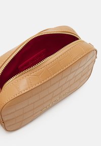 Tan läder kosmetikväska med präglat krokodilsmönster, guldzipper och burgundy innerfoder, med en diskret logotyp vid basen.