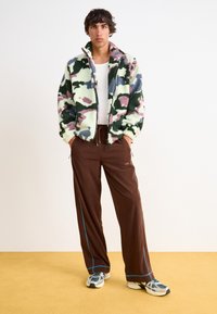 Fleecejacka med ett kamouflagemönster i grönt, rosa och blått, i kombination med bruna byxor med blå piping och snedställda fickor.