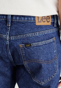 Pantalones de mezclilla azul con bolsillo trasero y etiqueta de la marca Lee, con costuras naranjas contrastantes, combinados con una blusa blanca.