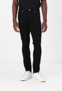 Hombre vestido con jeans negros ajustados y zapatillas blancas, de pie contra un fondo blanco liso.
