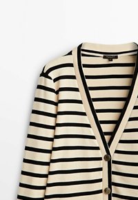 Cardigan beige avec des rayures horizontales noires, bordure noire le long du col en V et de la ligne de boutons, avec deux boutons ronds dorés.