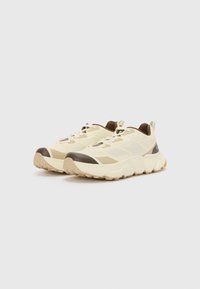 Beige Laufschuhe mit einem strukturierten Mesh-Obermaterial, dunkelbraunen Akzenten und einer klobigen Sohle. Mit runder Zehenpartie und kontrastierenden Schnürsenkeln.
