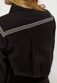 Čierna košeľa s plisovaným dizajnom na zadnej strane a bielym dekoratívnym prešívaním cez ramená. Vyrobená z textúrovaného materiálu.