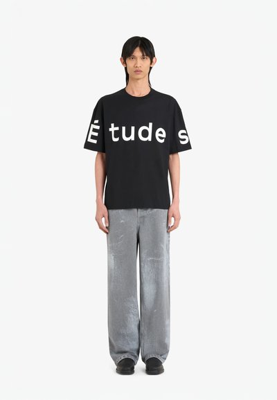 T-shirt noir avec un grand texte blanc "Études" sur le devant, associé à un pantalon gris clair ample. Le mannequin se tient droit, les mains sur les côtés.