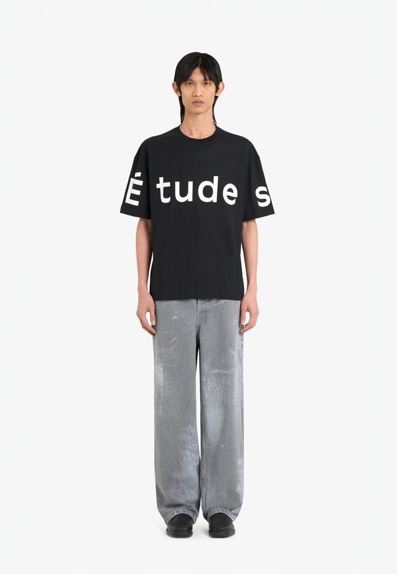 T-shirt noir avec un grand texte blanc "Études" sur le devant, associé à un pantalon gris clair ample. Le mannequin se tient droit, les mains sur les côtés.