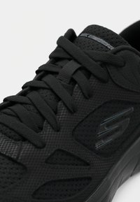 Sapatilha desportiva preta com parte superior texturizada em material sintético, detalhes em malha, atacadores planos e logo proeminente na lateral. Design elegante e moderno.