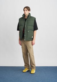 Roheline polsterdatud vest kõrge kaelusega, esipaneelil nööbid, küljetaskud, kantud musta T-särgi peale ja koos beezide tööpükste ning kollaste tossudega.