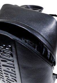 Mochila negra con una superficie texturizada, que presenta un gran logotipo en relieve en la parte frontal y un forro interior brillante. Cierre de cremallera visible.