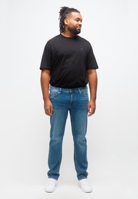 Άνδρας με ντραντέξ, φοράει μαύρο κοντομάνικο πουκάμισο, μπλε τζιν και λευκά αθλητικά παπούτσια, στέκεται μπροστά σε απλό λευκό φόντο.