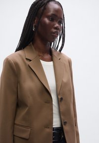 Brun blazer med et struktureret design, to knapper og hakede revers. Bruges over en hvid ribbet top, kombineret med et mørkt bælte.