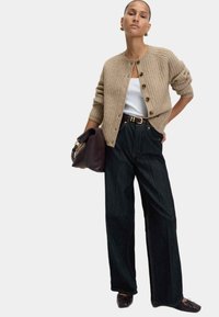 Cárdigan de punto beige con textura y botones, usado sobre una blusa blanca. Jeans anchos oscuros y un bolso oscuro completan el atuendo.