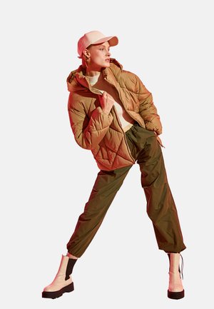 Frau lehnt seitlich und trägt eine beige gesteppte Jacke, einen weißen Pullover, olivfarbene Hose, weiße Stiefel und eine hellrosa Baseballkappe.