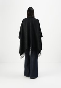 Persoon met steil, donker haar, gekleed in een zwarte franjesjaal en donkere wijde jeans, staand tegen een effen witte achtergrond, van achteren gezien.