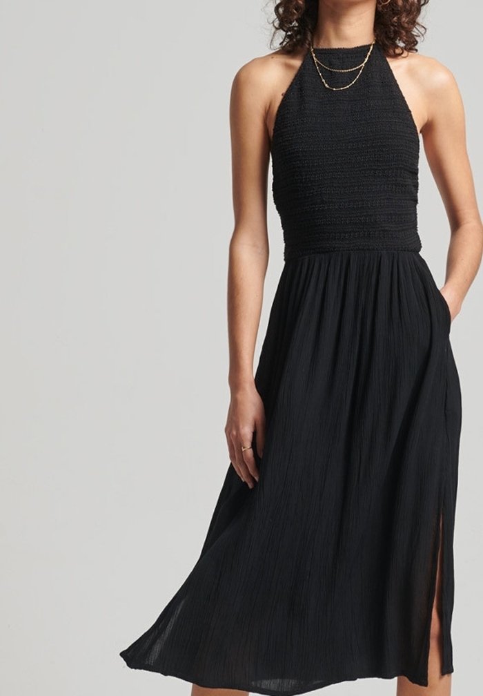 Femme portant une robe midi noire sans manches avec un corsage texturé, une jupe fluide, une fente latérale et des colliers dorés superposés sur un fond uni.