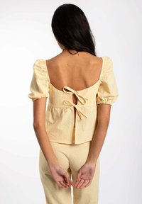 Haut à carreaux jaunes avec des manches bouffantes, un dos en scoop et une fermeture par nœud. Le tissu a un tissage texturé, présentant une taille cintrée avec un bas évasé.