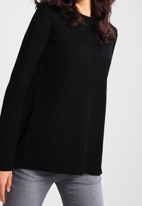 Musta ribbiknitisvuori, jossa on pikad varrukad ja lõdvestunud lõige, koos heledate hallide teksadega. Kangas näeb välja pehme ja tekstuurne.