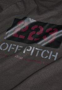 Dunkelgrauer Stoff mit einer glatten Textur zeigt einen großen Grafikdruck. Das Design umfasst "223" in Kastanienbraun, "OFF PITCH" in Silber und darunter Koordinaten.